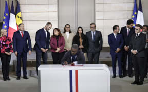 Member of the Palika (Kanak Liberation party) and L'UNI (National Union for Independance) coalition Jean-Pierre Djaiwe (C) signs the Elysee-Oudino agreement in front of French President Emmanuel Macron (2R) at the Elysee Palace in Paris on January 19, 2026. (Photo by STEPHANE DE SAKUTIN / POOL / AFP)