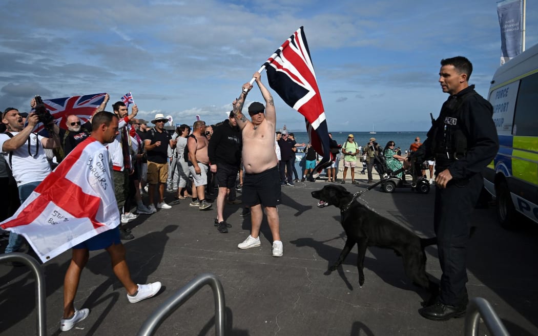 A police officer stands on duty as people wave Union Jacks and St George's flags during a 'Enough is Enough' demonstration called by far-right activists in Weymouth, on the southwest coast of England where the Bibby Stockholm migrant accommodation barge is moored, on August 4, 2024. Far-right protesters clashed with British police during tense rallies as unrest linked to disinformation about a mass stabbing that killed three young girls spread across the UK. (Photo by JUSTIN TALLIS / AFP)