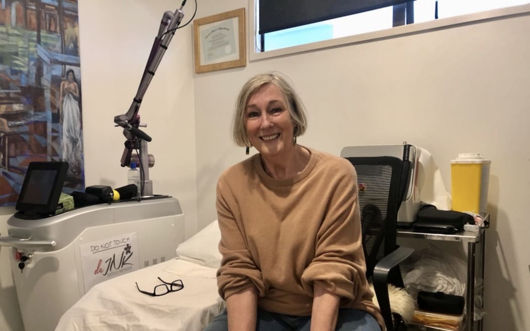 Helena sits in a tattoo removal clinic room. She sits next to a raised bed covered in a white sheet. There's a head lamp and some equipment next to the bench.