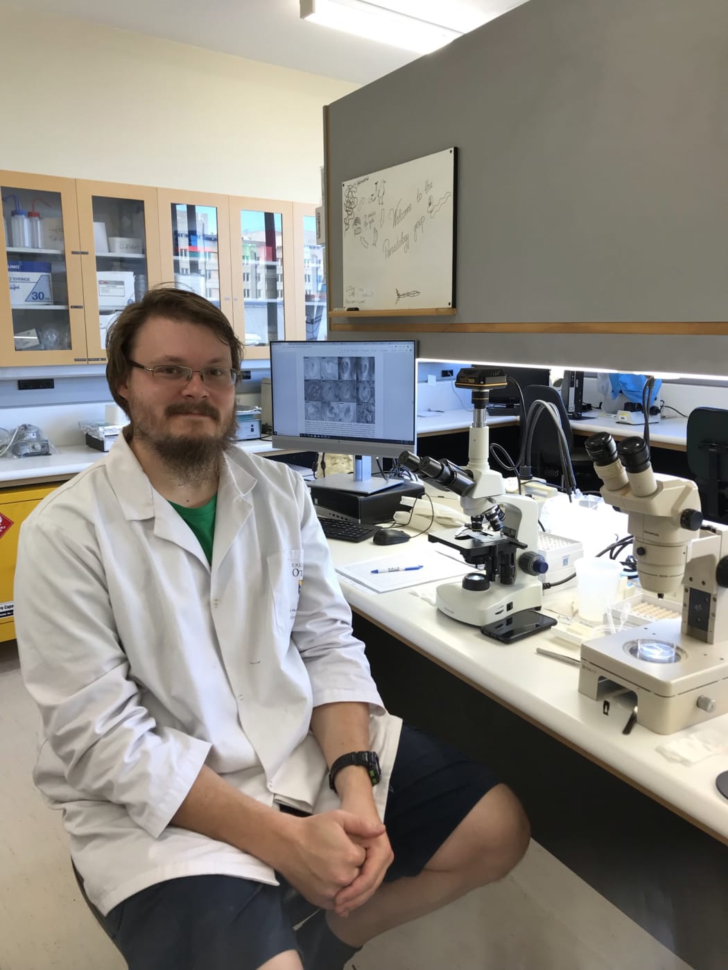 Jeff Doherty sits at a lab bench. In front of him there are two microscopes and a computer monitor. There are small plastic tubes, glass slides and a notebook on the lab bench beside the microscopes.