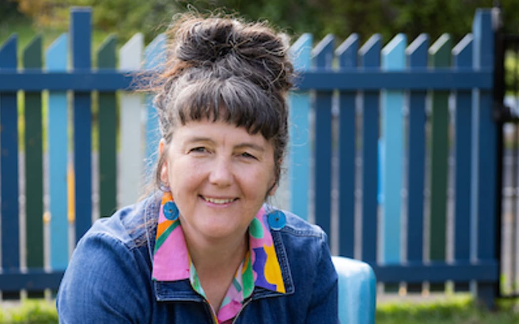 Colville School principal Susie Sumner is seated in an outdoor chair in the school grounds. She wears a denim jumpsuit over a colourful shirt and is patting a black-and-white dog.
