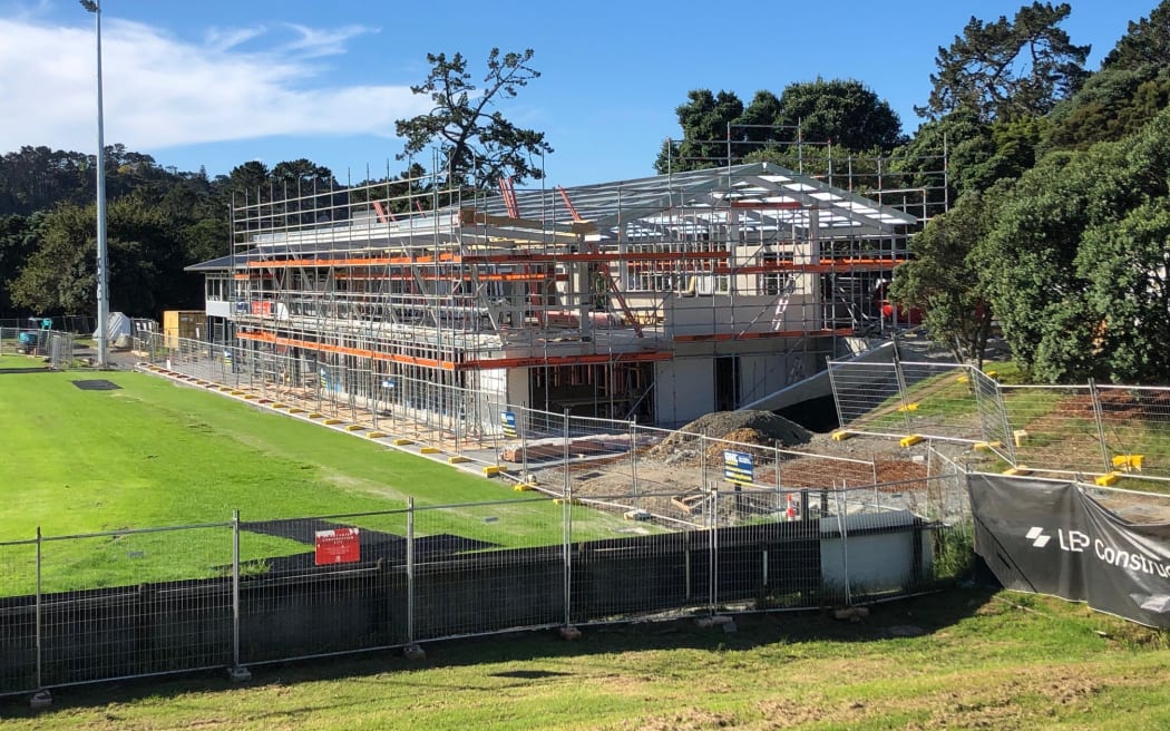 A large building is being constructed next to a green sports field. The structure is covered in scaffolding.