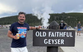 Ash Jurberg is holding a can of Yellowstone branded beer and standing next to a sign that says "Old Faithful Geyser". In the background you can see steam coming off the geyser.