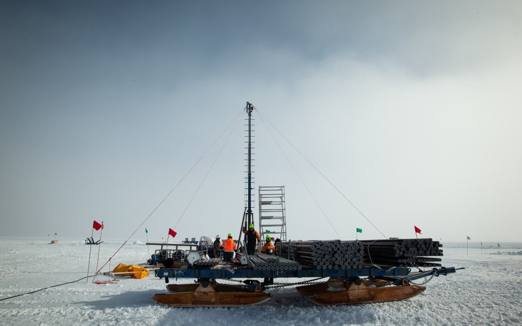 A group of people assemble pieces of metal piping, on top of a very large sled, against the snow and ice of Antarctica with grey clouds gathering in the background.