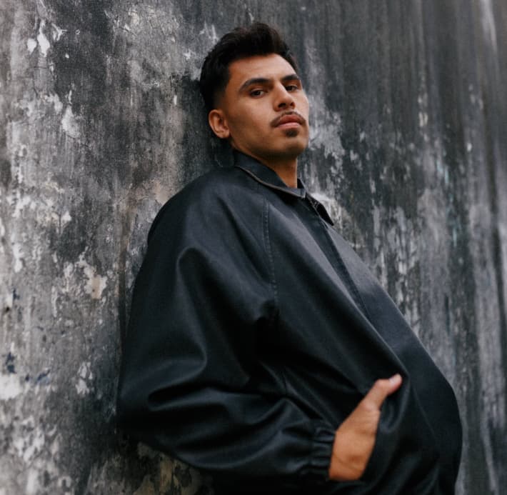 MOHI leans against a paint-splattered wall in a black leather jacket.