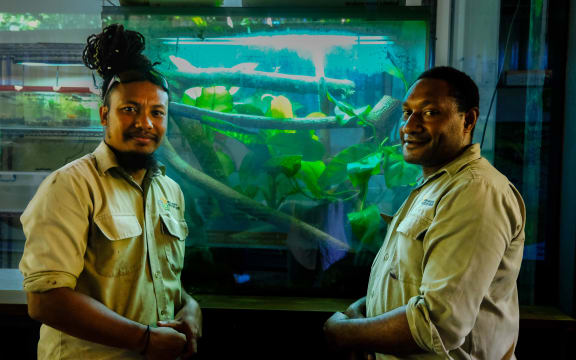 Ryan and his colleague, from Papua New Guinea, stand in front of a tank filled with greenery. They are both wearing khaki shirts.