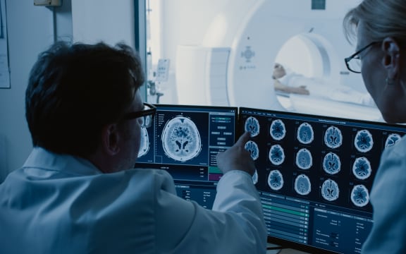 In Control Room Doctor and Radiologist Discuss Diagnosis while Watching Procedure and Monitors Showing Brain Scans Results, In the Background Patient Undergoes MRI or CT Scan Procedure.