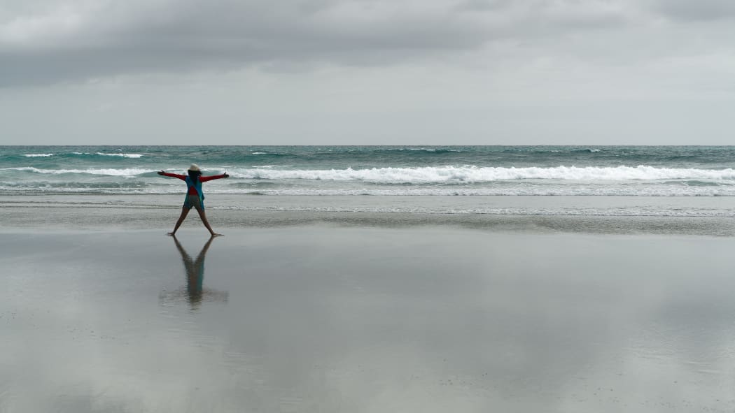 Girl, back to camera, doing exercises stretching arms outstretched on an empty beach, New Zealand.