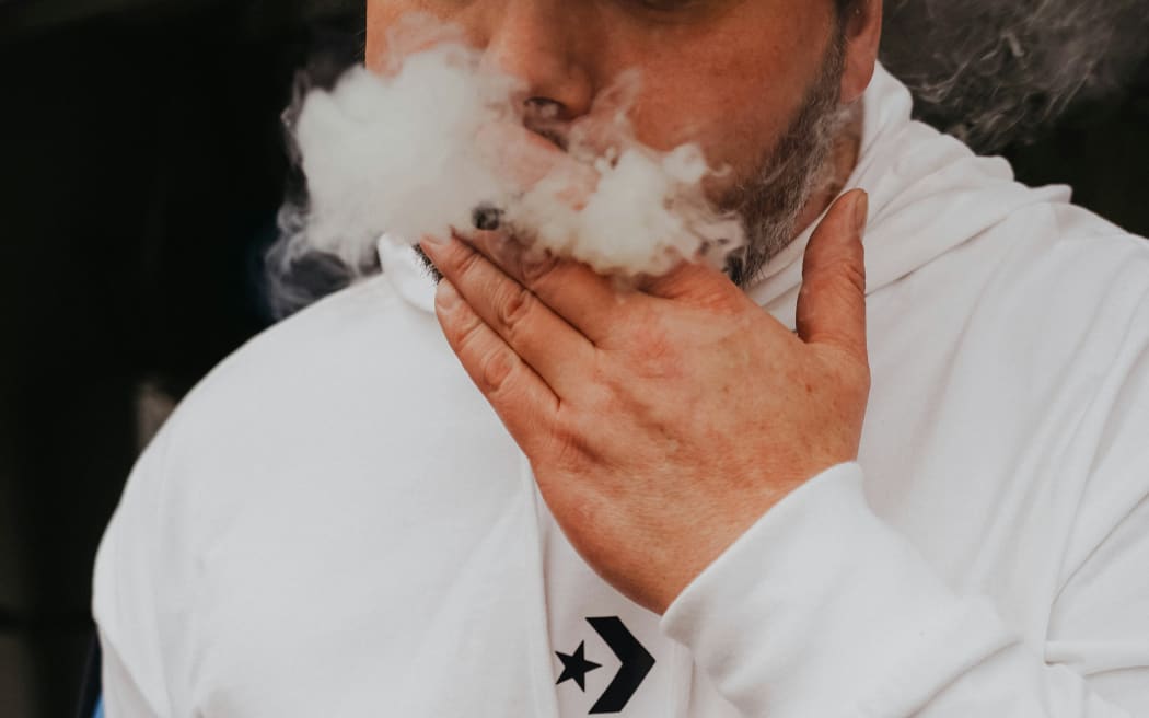 A bearded man in a hoodie inhales smoke.