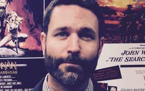 Andrew looks at the camera. He is standing in front of a wall of movie posters. He wears a collared checked shirt and a black jacket.