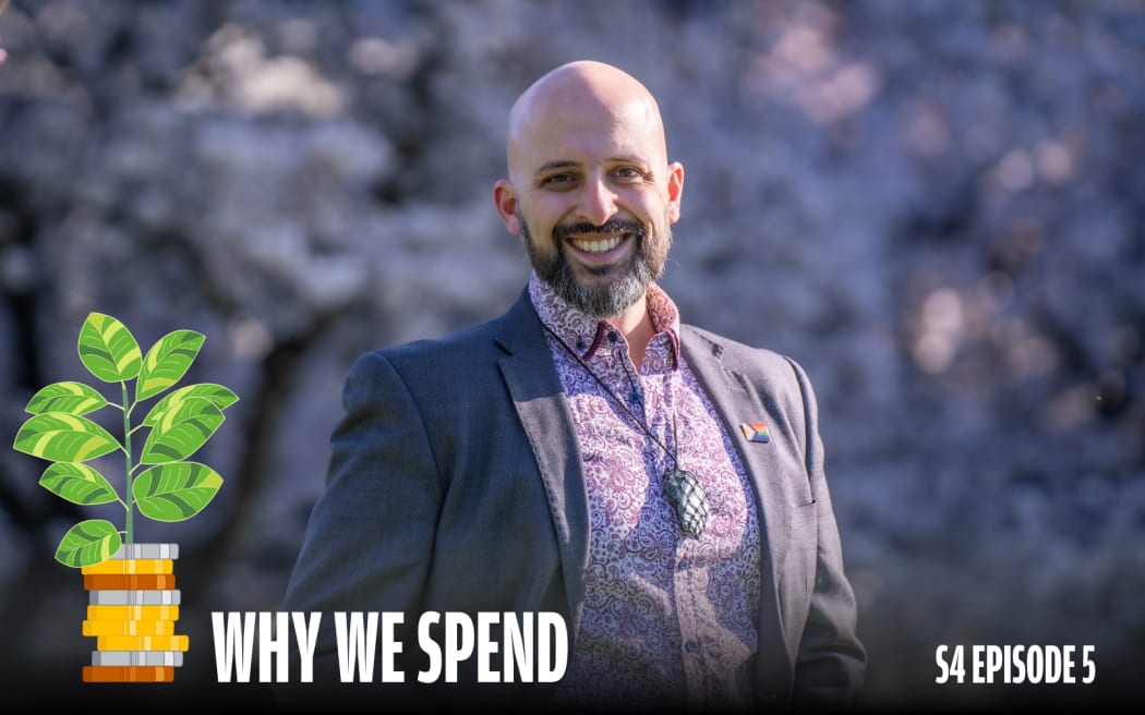 Smiling man in a patterned shirt and dark blazer stands outdoors in front of blurred purple foliage. He wears a pendant and a rainbow pin on his lapel. A cartoon plant growing out of stacked coins appears beside him, with bold white text reading 'Why We Spend' and smaller text in the corner: 'S4 Episode 5'