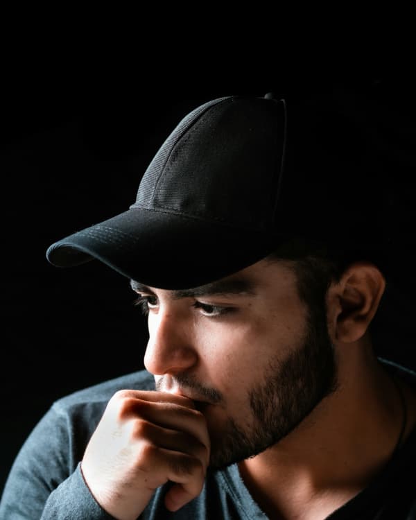 A nervous bearded man in a cap holds his chin.