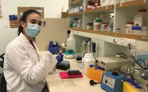 Lucy sits at a lab bench with a pipette in hand. There is a tray of small tubes in front of her as well as an array of different lab equipment.