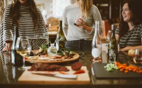 People stand around a table with food on it, some wine bottles are on the table, some people are holding glasses of drink.