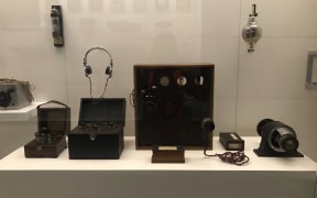 Shows a display case with some early radio equipment used by Professor Robert Jack. A large wooden framed transmitter sits in the centre with a generator and microphone to the side.