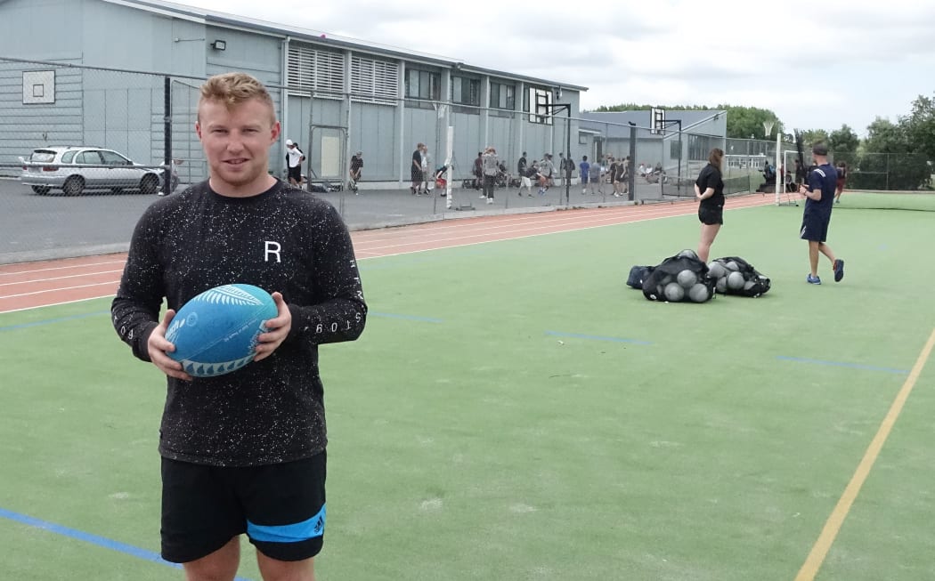A young teacher stands on a field of astro turf with a rugby ball in hand