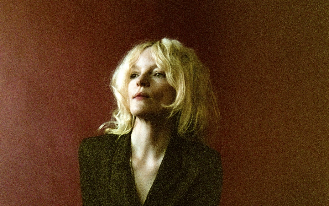 A woman with blonde hair in a black suit poses in front of a red background.
