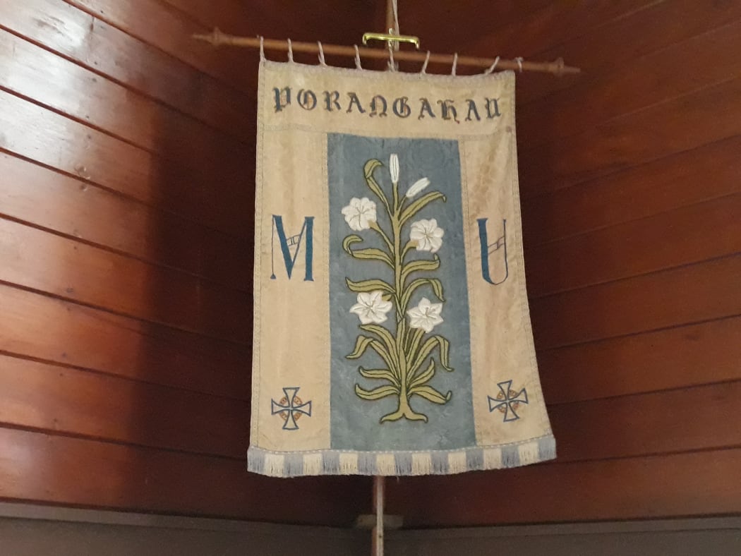A fabric banner with a stem of white lilies sewn on is displayed in a corner at the front of St Michael and All Angels Church, Porangahau.