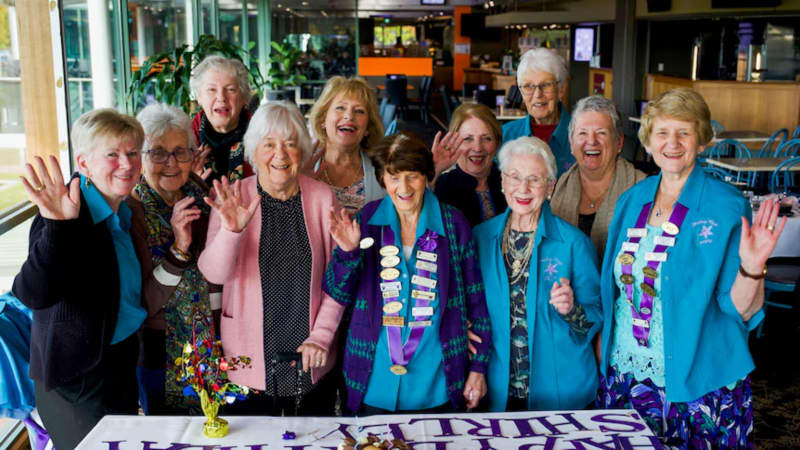A group of 11 women from the ACT Shirley Club stand waving at the camera.