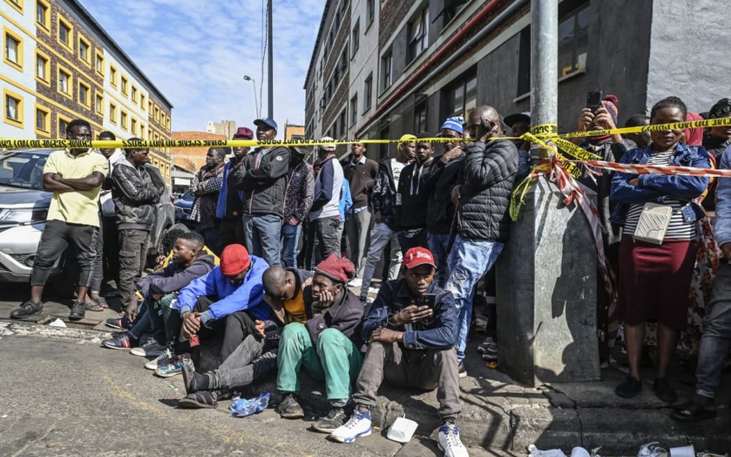 Residents, relatives of victims and members of the public gather behind a police tape at the site of a burned apartment block in Johannesburg on 1 September, 2023.
