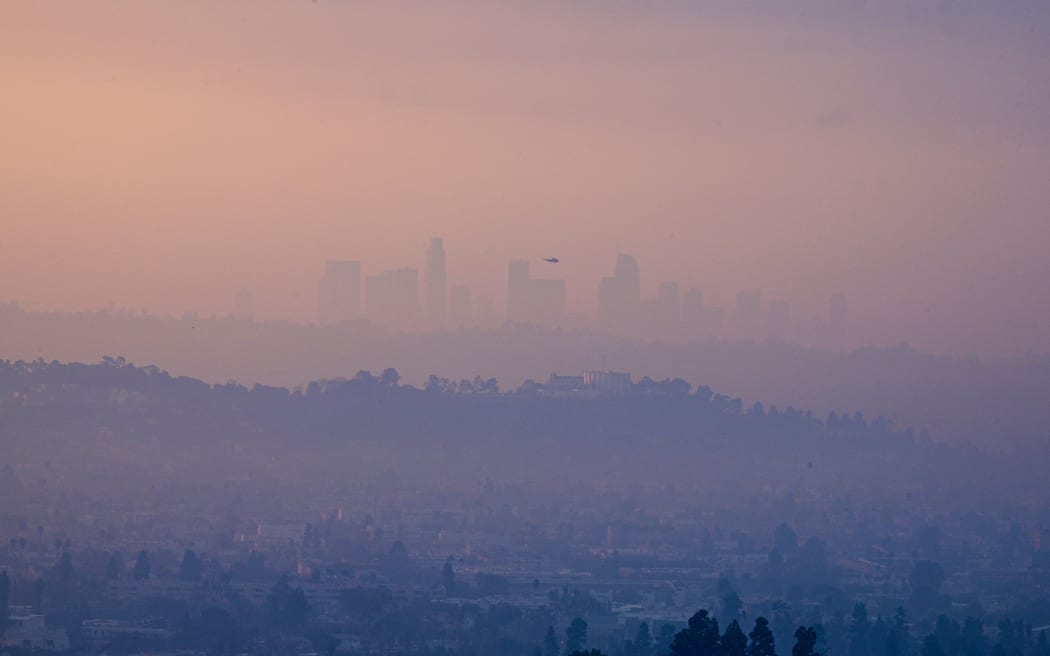 A helicopter flies past downtown Los Angeles, California, covered in smoke from the Eaton Fire, Palisades Fire and other smaller fires, as seen from Glendale, California, on January 9, 2025. Wildfires threatened to engulf parts of Hollywood on January 9 as a growing number of blazes raged across Los Angeles, forcing over 100,000 people to flee their homes and claiming at least five lives. (Photo by Robyn Beck / AFP)