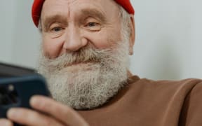 A senior man with a beard and red beanie smiles at his phone.