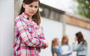 A photo of a sad looking teenage girl in the foreground. Behind her is a group of other girls talking