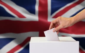 Close-up of man casting and inserting a vote and choosing and making a decision what he wants in polling box with United Kingdom or UK flag blended in background.