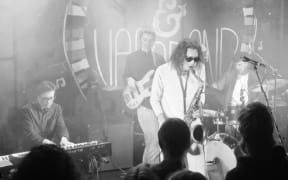 A black and white image of musicians playing keyboard, bass guitar, saxophone, and drums