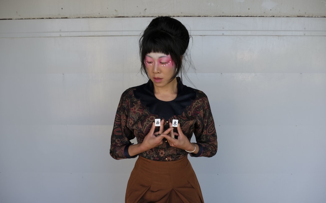 A woman in a brown shirt with a black collar stands in front of a white wall. She holds up two white cubes inscribed with Chinese characters.