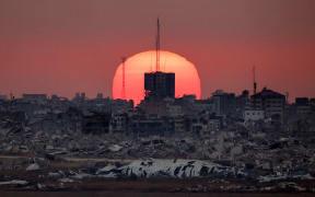 This picture taken from a position on the Israeli border with the Gaza Strip, shows the sun setting behind destroyed buildings in the Palestinian territory, on July 1, 2025. (Photo by Jack GUEZ / AFP)