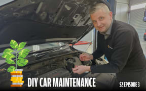 A man is standing in front of a car with its bonnet up. He is holding out the oil dipstick.