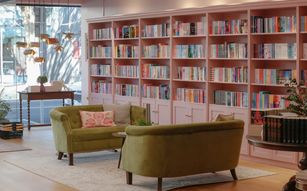The interior of Enamoured Books in Remuera. Two plush green couches face each other in the shop floor, while the walls are lined with pink shelves full of brightly-coloured book spines.