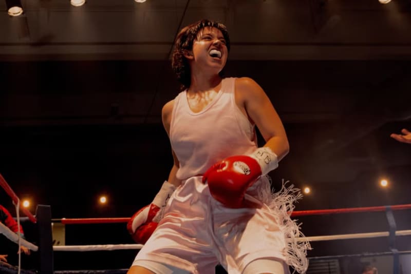 Sydney Sweeney has short dark hair and stands in a boxing ring wearing boxing gloves and a mouthguard.