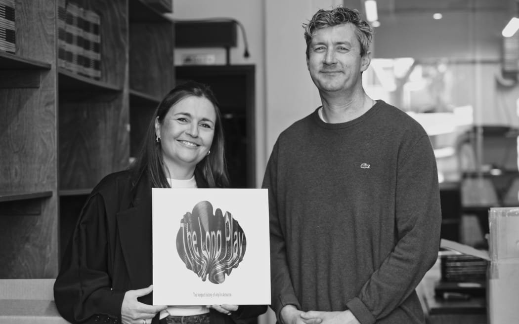 This is a black and white photo of Charlotte Ryan and Duncan Greive holding a vinyl copy of their new podcast.