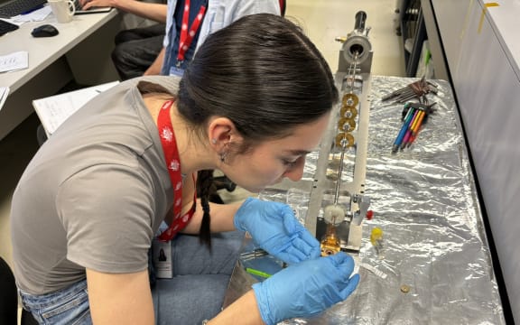A woman wearing blue gloves carefully handles a small piece of metal at the end of a long rod-shaped contraption sitting on a bench covered in tinfoil.