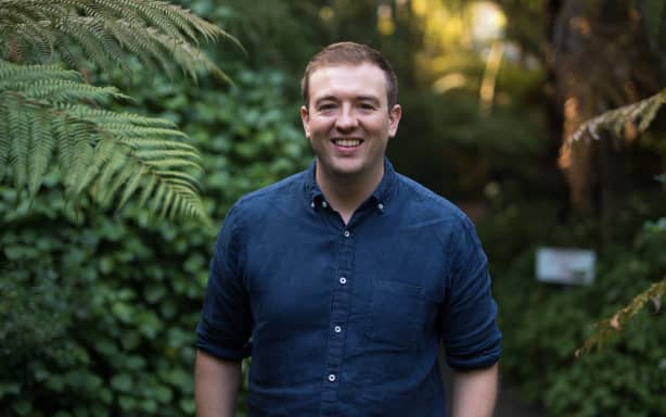 A portrait shot of Luke from the waist up. He is smiling, and wearing a blue shirt. Some ferns and other greener is visible beside and behind him.
