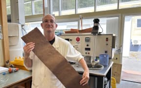 Sean is standing in front of a small scale paper making machine, that looks a bit like a top-loader washing machine. He is wearing glasses and a long white lab coat, and is holding a long strip of brown parchment across his torso.