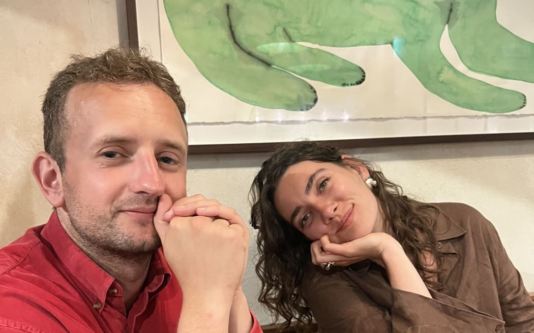 Gregor is on the right in a red shit. There is a woman (possibly his partner) sat on the left. They appear to be in a cafe or resturant.