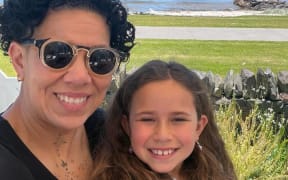 A woman with short hair and a child with long hair smile and sit close together with a coastal view behind them.
