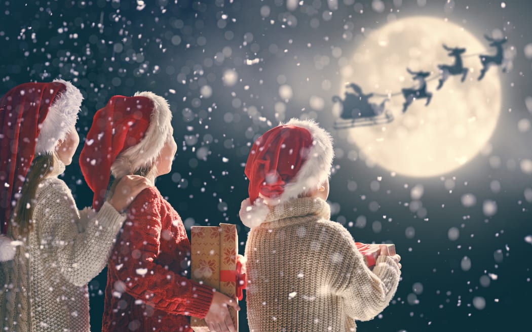 Merry Christmas and happy holidays! Cute little children with xmas presents. Santa Claus flying in his sleigh against moon sky. Kids enjoying the holiday with gifts on dark background.