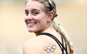 A photograph of Olivia Podmore, looking back at the photographer over her shoulder and smiling. She wears a black singlet and has a tattoo of the Olympic rings on her shoulderblade.