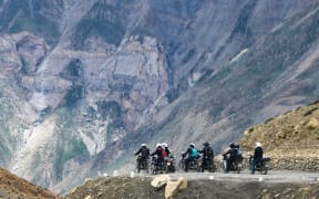A group of motorcycle riders are on a small road halfway up a mountain. The mountain towers behind them.