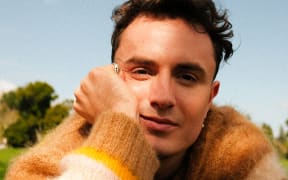 Cammy smiles at the camera. He is lying on his stomach on the grass with his head propped up in his hand. He wears a fluffy yellow jumper.
