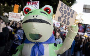 A person in an inflatable frog suit holds a sign during a protest in Los Angeles, California on January 10, 2026 against US Immigration and Customs Enforcement (ICE) after the fatal shooting of Renee Nicole Good in Minneapolis. A US Immigration and Customs Enforcement (ICE) agent shot and killed 37-year-old Renee Nicole Good on the streets of Minneapolis on January 7, leading to huge protests and outrage from local leaders who rejected White House claims she was a domestic terrorist. (Photo by ETIENNE LAURENT / AFP)