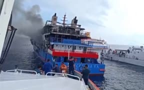 Dozens of people can be seen floating in the ocean, watching the KM Barcelona 5 ferry as it burns.
