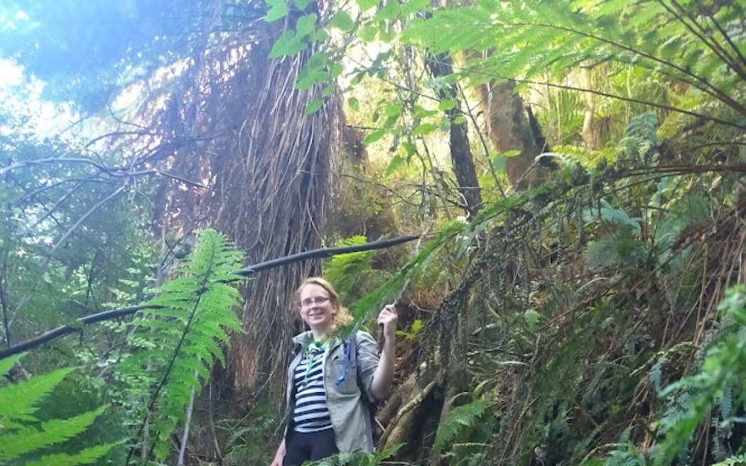 Luna standing in the bush surrounded by ferns. She is wearing a rain jacked and backpack.