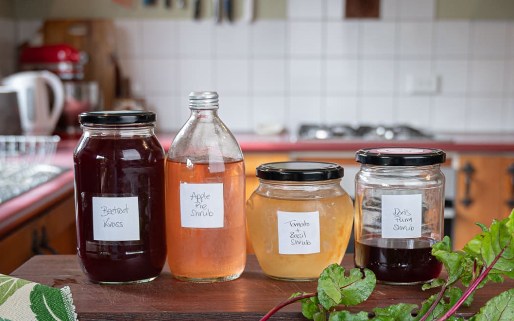 An array of different 'Shrubs' drinks made out of left over veggies, fruits and herbs, in glass jars, lined up on a wooden bench. A beetroot sits in the foreground.