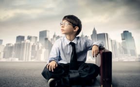 A boy dress in a suit and leaning on a suitcase in front of tall office buildings contemplates the future of work.