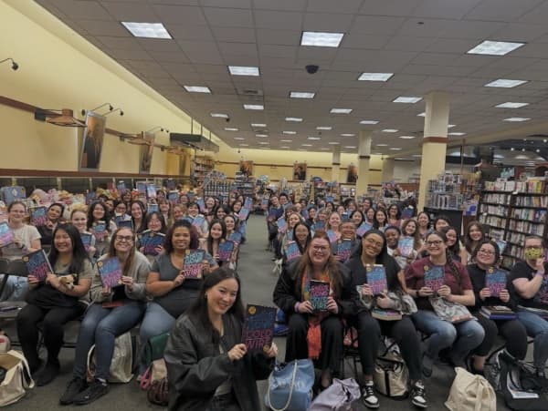 Chloe Gong on her US book tour holds up a copy of her latest book, Cold Wire, as she sits between dozens of fans holding her book in a hall.